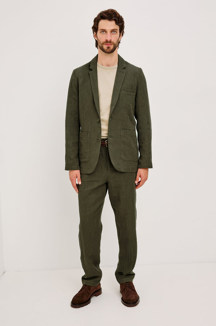 The Bond is a single breasted blazer with two buttons, one chest pocket and two patch pockets in Deep Olive colorway. Fullbody front view featured on model.