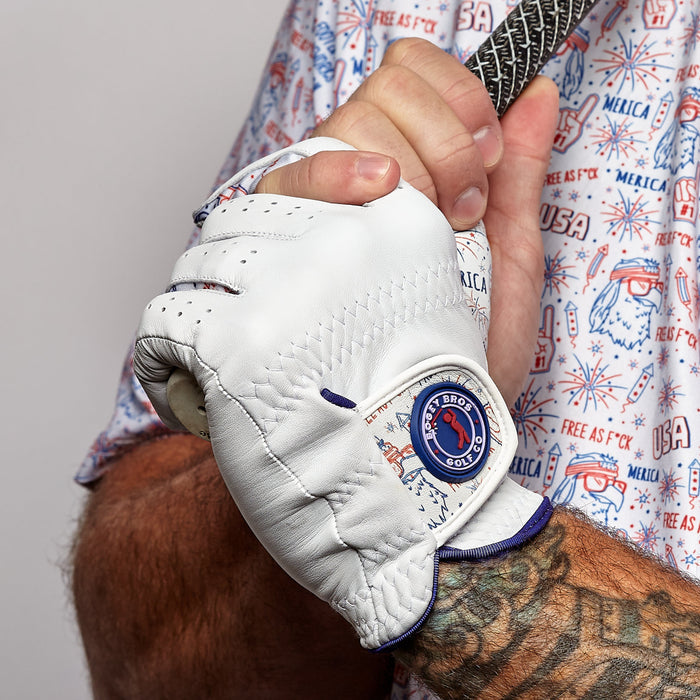 A tattooed arm grips a golf club, wearing the 'Merica - Glove: a precision-fit Cabretta leather glove with a blue and red Palm Bird Golf patch. The shirt displays cartoon USA motifs, sunglasses, eagles, and “MERICA” in red and blue.