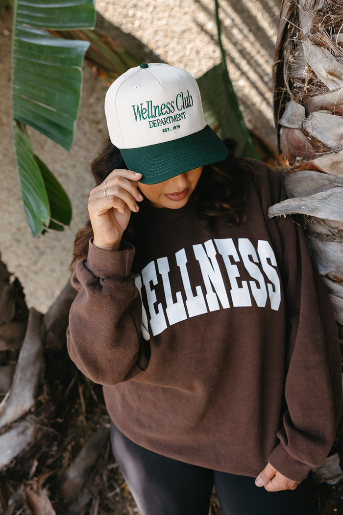 Person wearing a Wellness Club Department Hat | Lucky Owl with green and white colors, paired with a brown sweatshirt featuring large wellness text, standing near tropical foliage.