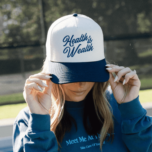 Woman wearing a white and navy blue "Health Is Wealth Hat | Lucky Owl" adjusting the brim outdoors with blurred green foliage in the background.