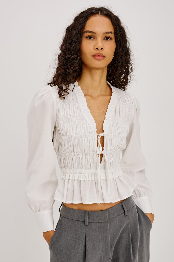 Sami Shirt in Cotton Poplin With V-Neckline Front Ties, Shirred Fitted Bodice, Flared Hem, Ruffle Trim, and Long Puff Sleeves With Buttoned Cuffs in White Colorway - Upper Body Front View Featured on Model