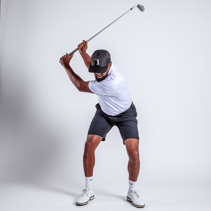 Wearing the Stealth Mode - Light - Polo (white, UPF 50), black shorts, white socks, shoes, and a black cap, a man prepares to swing a golf club indoors. He is at the top of his backswing on a plain white background.