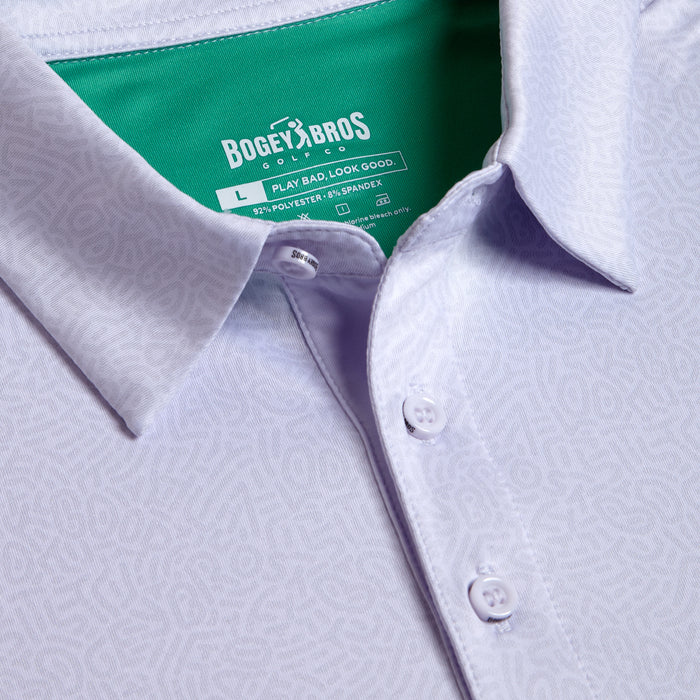 Close-up of the Stealth Mode - Light - Polo, a light purple collared shirt with a subtle geometric pattern. It features moisture-wicking fabric, white buttons, and a green inner collar tag reading BOGEY BROS with size L and fabric details in white.
