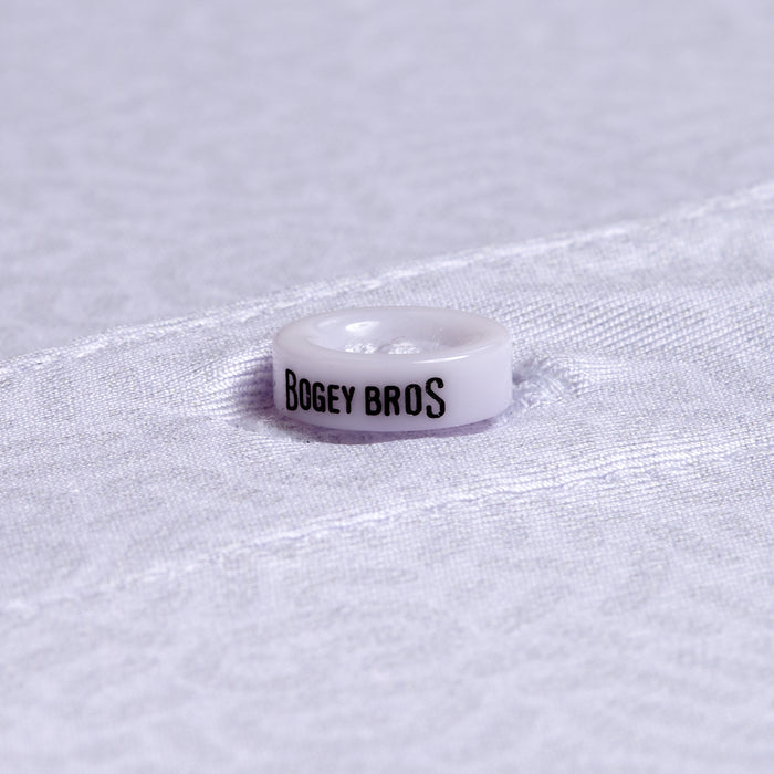 A close-up of a white button labeled BOGEY BROS in black caps on the Stealth Mode - Light - Polo’s moisture-wicking white, subtly textured fabric, with an athletic fit and a horizontal seam by the button.